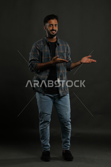Gestures of affirmation and identification of a specific place with expressions of pleasure, portrait of a Saudi Gulf Arab man wearing casual clothing raising his hands to indicate something, confidence in knowing something, full-length body portrait, black background
