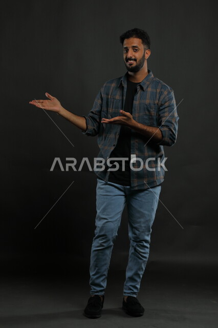 Gestures of affirmation and identification of a specific place with expressions of pleasure, portrait of a Saudi Gulf Arab man wearing casual clothing raising his hands to indicate something, confidence in knowing something, full-length body portrait, black background