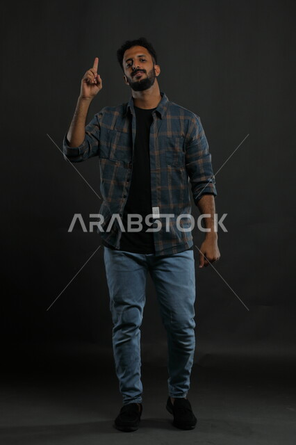 التهديد والتوعد لشيء ما، ايماءات الاستفسار والاشارة للاعلى , الوقوف باستقامة والنظر إلى الكاميرا, بورتريه لرجل عربي خليجي سعودي مبتسم يرتدي الزي الكاجوال يرفع اصبع السبابة بإيماءات التوضيح, صورة الجسم بالطول الكامل , خلفية سوداء