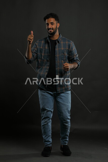 النظر إلى الكاميرا بايماءات القبول والموافقه, الشعور بالإعجاب والرضا والسعاده، بورتريه لشاب وسيم عربي خليجي سعودي مبتسم يرتدي الزي الكاجوال ويرفع أصبع الابهام بعلامة الجودة ،صورة الجسم بالطول الكامل، خلفية سوداء