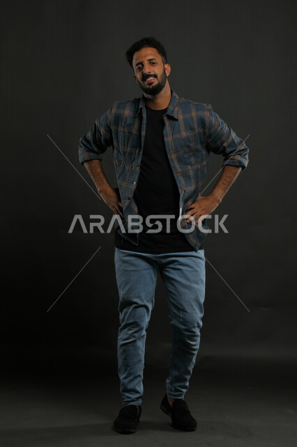 Asking and inquiring about something, a portrait of a Saudi Gulf Arab man wearing casual clothes and placing his hands on his waist with gestures of bewilderment and amazement, making fashion presentations with expressions of pleasure, full-length body portrait, black background.