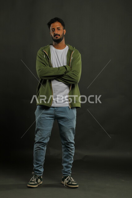 النظر إلى الكاميرا بإيماءات أيدي متقاطعه، بورتريه لشاب وسيم عربي خليجي سعودي يرتدي الزي الكاجوال يقف باستقامة وثقه بالنفس، مفهوم الاناقه و الاهتمام بالمظهر الخارجي، صورة الجسم بالطول الكامل، خلفية سوداء
