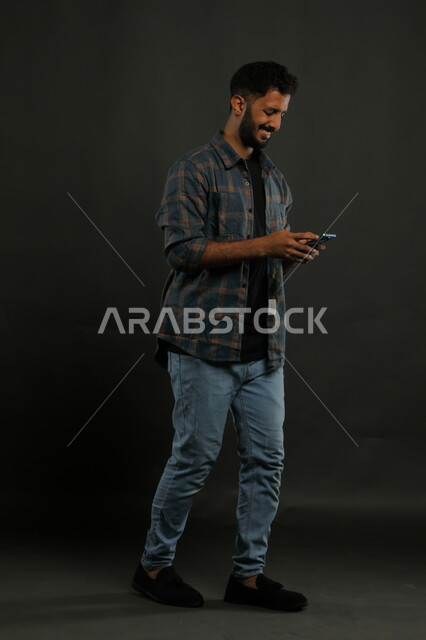تصفح تطبيقات ومواقع التواصل الاجتماعي، استخدام الأجهزة والتقنيات الحديثة، محادثات ودردشات مع الأصدقاء, بورتريه لشاب عربي سعودي خليجي مبتسم يرتدي الزي الكاجوال يمشي يمسك الهاتف المحمول, إيماءات السعادة والفرح, صورة الجسم بالكامل, خلفية سوداء.