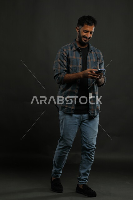 Using modern devices and technologies, conversations and chats with friends, browsing social networking applications and websites, a portrait of a smiling Saudi Arab Gulf man wearing a casual outfit, walking and holding a mobile phone, gestures of happiness and joy, full body image, black background