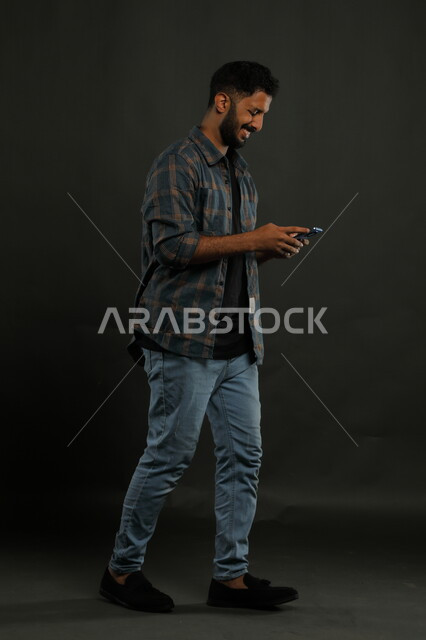 محادثات ودردشات مع الأصدقاء, تصفح تطبيقات ومواقع التواصل الاجتماعي، بورتريه لشاب عربي سعودي خليجي مبتسم يرتدي الزي الكاجوال يمشي يستخدم الهاتف المحمول, إيماءات السعادة والفرح, صورة الجسم بالكامل, خلفية سوداء.