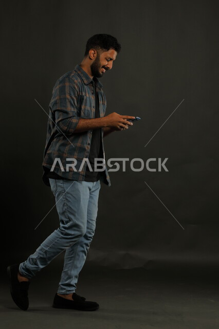 تصفح تطبيقات ومواقع التواصل الاجتماعي، محادثات ودردشات مع الأصدقاء, بورتريه لشاب عربي سعودي خليجي مبتسم يرتدي الزي الكاجوال يمشي يستخدم الهاتف المحمول, إيماءات السعادة والفرح, صورة الجسم بالكامل, خلفية سوداء.