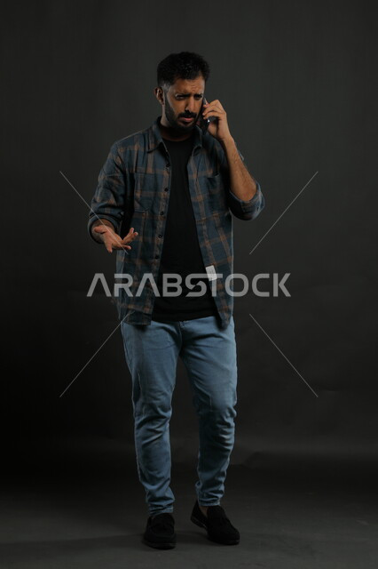إيماءات الغضب والقلق حول موضوع معين، التساؤل والاستفهام عن أمر ما، استخدام الأجهزة والتقنيات الحديثة، جدالات ونقاشات عبر الجوال, بورتريه لشاب عربي سعودي خليجي يرتدي الزي الكاجوال يمسك الهاتف المحمول, صورة الجسم بالكامل, خلفية سوداء.