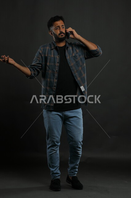 التساؤل عن أمر معين،  الشعور بالجهل وعدم المعرفة, بورتريه لشاب عربي سعودي خليجي يرتدي الزي الكاجوال يستخدم الهاتف المحمول يرفع يده بإيماءات الاستفسار والاستفهام, التواصل مع الأهل والأصدقاء, صورة الجسم بالكامل, خلفية سوداء.