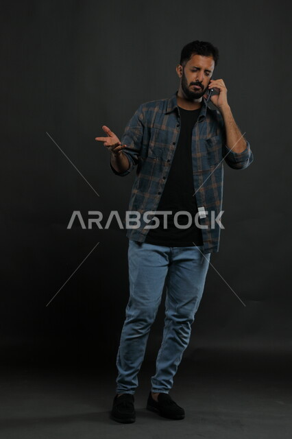  الشعور بالجهل وعدم المعرفة, بورتريه لشاب عربي سعودي خليجي يرتدي الزي الكاجوال يستخدم الهاتف المحمول يرفع يده بإيماءات الاستفسار والاستفهام, التساؤل عن أمر معين، التواصل مع الأهل والأصدقاء, صورة الجسم بالكامل, خلفية سوداء.