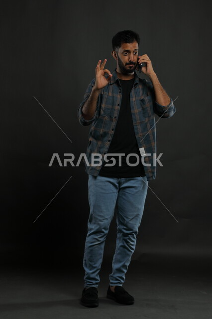 إيماءات الفرح والرضا بسماع الأخبار السارة, الإعجاب بالتقنيات الحديثة, بورتريه لشاب عربي سعودي خليجي يرتدي الثوب والشماغ التقليدي مبتسم يستخدم الهاتف المحمول ينظر إلى الكاميرا, صورة الجسم بالكامل, خلفية سوداء.