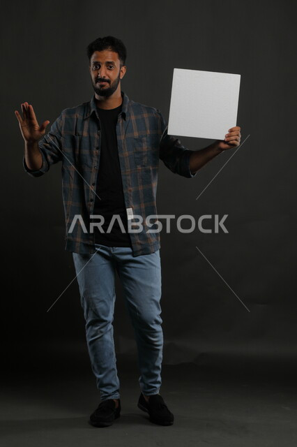إعلانات وعروض تسويقية، موك اب مربع أبيض، استخدام لوحات توضيحية ووسائل تعليمية، بورتريه لشاب عربي خليجي سعودي مبتسم يرتدي الزي الكاجوال يمسك بيده لوحه بيضاء مربعه فارغة وينظر إلى الكاميرا، صورة الجسم بالكامل، خلفية سوداء