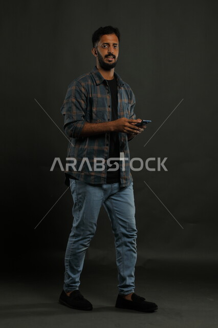 تصفح برامج وتطبيقات التواصل الاجتماعي، استخدام الأجهزة والتقنيات الحديثة, بورتريه لشاب عربي سعودي خليجي يرتدي الزي الكاجوال مبتسم ينظر إلى الكاميرا يستخدم الهاتف المحمول، صورة الجسم بالكامل, خلفية سوداء.