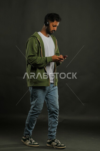 تبادل الاحاديث مع الاصدقاء عبر تطبيقات الجوال أثناء الاستماع إلى المقاطع الموسيقيه المفضلة، بورتريه لشاب وسيم عربي خليجي سعودي يرتدي الزي الكاجوال يحمل بيده الهاتف المحمول ويضع على رأسه سماعات الهيدفون ، صورة الجسم بالكامل، خلفية سوداء