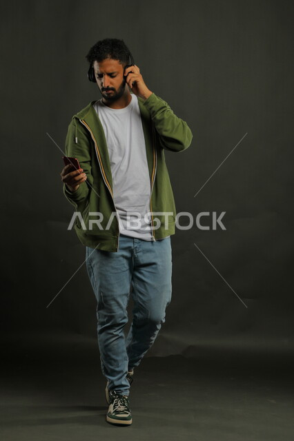 استخدام تطبيقات الاغاني والتسجيلات الصوتية في الجوال، الدردشة والتحدث مع الأصدقاء عبر تطبيقات الهاتف المحمول، بورتريه لشاب عربي سعودي خليجي يرتدي الزي الكاجوال يضع سماعات الهيدفون، صورة الجسم بطول الكامل، خلفية سوداء