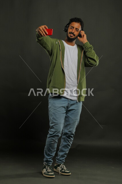 وضع سماعات الهيدفون الحديثة بإيماءات السرور، الاستمتاع بسماع المقاطع الموسيقية والصوتية،  بورتريه لشاب عربي خليجي سعودي يرتدي الزي الكاجوال يقوم بتوثيق اللحظات السعيدة بالتقاط صور السيلفي عبر الجوال، صورة الجسم بالكامل، خلفية سوداء