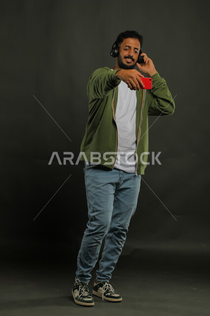 توثيق اللحظات السعيدة بالتقاط صور السيلفي عبر الجوال، الاستماع إلى المقاطع الموسيقيه المفضلة، بورتريه لشاب عربي خليجي سعودي يرتدي الزي الكاجوال يحمل بيده الهاتف المحمول ويضع سماعات الهيدفون الحديثة بإيماءات السرور، صورة الجسم بالكامل، خلفية سوداء