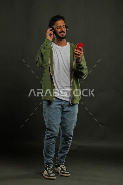 استخدام تقنية الهيدفون الحديثه، اختيار الأغاني بإيماءات السرور، الاستماع إلى المقاطع الموسيقيه المفضلة، بورتريه لشاب وسيم عربي خليجي سعودي يرتدي الزي الكاجوال يحمل بيده الجوال ويضع سماعة الرأس الصوتية ، صورة الجسم بالطول الكامل، خلفية سوداء