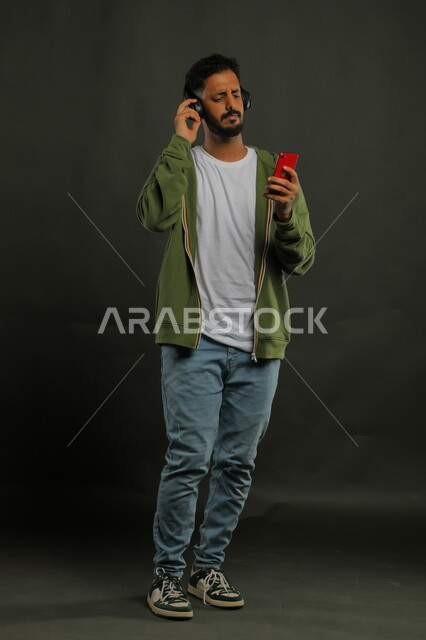 ايماءات الاندماج والاستمتاع، تطبيقات الاغاني والتسجيلات الصوتية في الهاتف المحمول، الاستماع إلى الأغاني عبر الهيدفون، بورتريه لشاب عربي خليجي سعودي يرتدي الزي الكاجوال وسماعة الرأس يحمل بيده الجوال، صورة الجسم بالطول الكامل، خلفية سوداء