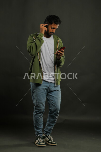 Exchanging conversations with friends via mobile applications while listening to favorite music clips, portrait of a handsome Saudi Gulf Arab young man wearing a casual outfit, holding a mobile phone in his hand and wearing headphones on his head, full body portrait, black background