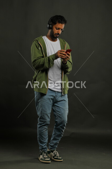 تبادل الاحاديث مع الاصدقاء عبر تطبيقات الجوال أثناء الاستماع إلى المقاطع الموسيقيه المفضلة، بورتريه لشاب وسيم عربي خليجي سعودي يرتدي الزي الكاجوال يحمل بيده الهاتف المحمول ويضع على رأسه سماعات الهيدفون ، صورة الجسم بالكامل، خلفية سوداء