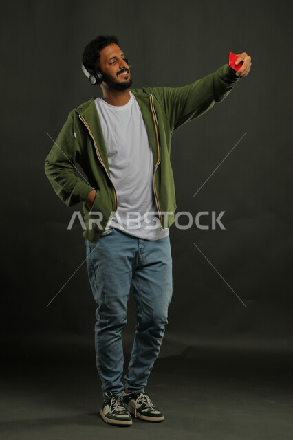 استخدام تقنية الهيدفون الحديثه بإيماءات السرور، التقاط صور السيلفي عبر الجوال، الاستماع إلى المقاطع الموسيقيه المفضلة، بورتريه لشاب عربي خليجي سعودي يرتدي الزي الكاجوال يحمل بيده الهاتف المحمول ويضع سماعة الرأس الصوتية، صورة الجسم بالكامل، خلفية سوداء