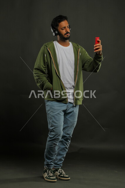 التحدث والدردشة مع الاصدقاء أثناء الاستماع إلى المقاطع الموسيقيه المفضلة، بورتريه لشاب وسيم عربي خليجي سعودي يرتدي الزي الكاجوال يحمل بيده الهاتف المحمول ويضع سماعة الرأس الصوتية ، استخدام تقنية الهيدفون الحديثه، صورة الجسم بالطول الكامل، خلفية سوداء