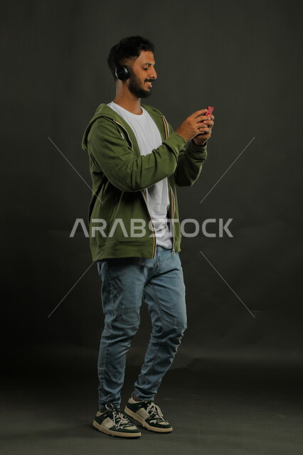 استخدام تقنية الهيدفون الحديثه، الاستماع إلى المقاطع الموسيقيه المفضلة بإيماءات السرور، بورتريه لشاب وسيم عربي خليجي سعودي يرتدي الزي الكاجوال يحمل بيده الهاتف المحمول ويضع سماعة الرأس الصوتية ، صورة الجسم بالطول الكامل، خلفية رماديه