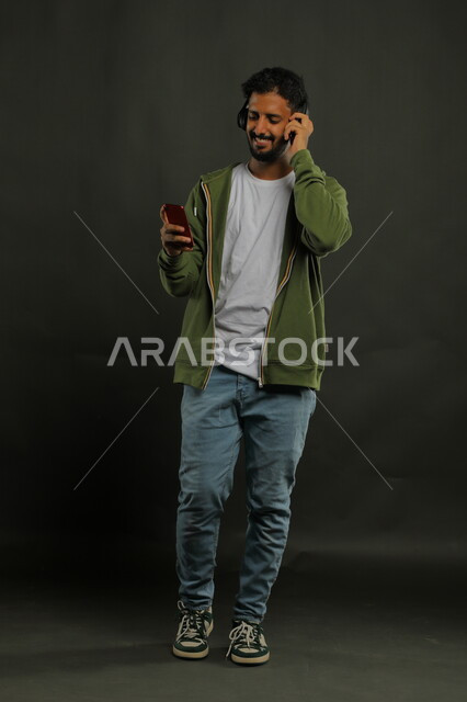 Gestures of pleasure and integration using a mobile phone, using song applications and audio recordings on mobile phones, advanced modern headphone technology, a portrait of a young Saudi Arab Gulf man wearing a casual outfit with a loudspeaker on his head, full body portrait, black background