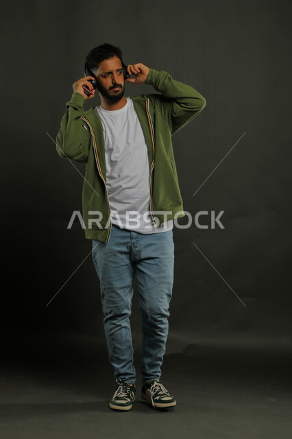 استخدام تقنية الهيدفوون الحديثة المتطورة، الاستماع إلى المقاطع الموسيقيه المفضلة، بورتريه لشاب عربي خليجي سعودي مبتسم يرتدي الزي الكاجوال ويضع سماعات الرأس, رفع اليدين إلى الأعلى لمسك مكبر الصوت بإيماءات الاندماج ، صورة الجسم بالطول الكامل، خلفية سوداء