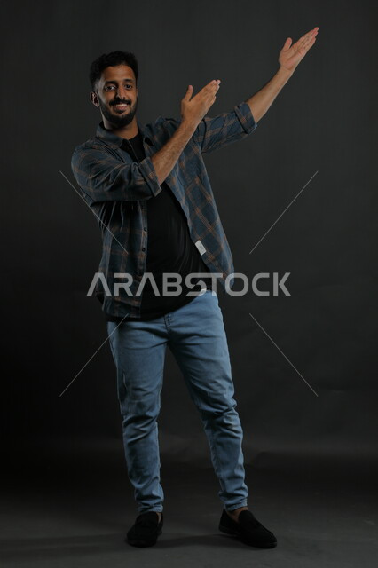 إشارات وحركات يدين تدل على هذا المكان (هنا)، النظر إلى الكاميرا ،تحديد موقع معين بإيماءات السرور والسعاده، بورتريه لرجل عربي خليجي سعودي يرتدي الزي الكاجوال يشير بيديه نحو الأعلى، صورة الجسم بالطول الكامل، خلفية سوداء.