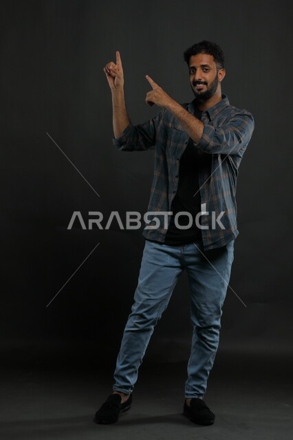 تحديد موقع معين بإيماءات السرور والسعاده، إشارات وحركات يدين تدل على هذا المكان (هنا)، بورتريه لرجل عربي خليجي سعودي يرتدي الزي الكاجوال يشير بأصابعه نحو الأعلى، صورة الجسم بالطول الكامل، خلفية رمادية.