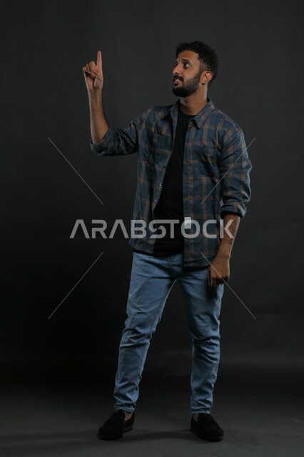 الوقوف باستقامة والنظر إلى شيء ما, ايماءات الاستفسار والاشارة إلى شيء معين, بورتريه لرجل عربي خليجي سعودي يرتدي الزي الكاجوال يرفع اصبعه للأعلى بإيماءات التوضيح, صورة الجسم بالطول الكامل , خلفية سوداء