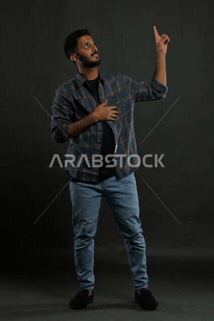 الوقوف باستقامة والنظر إلى شيء ما, ايماءات الاستفسار والاشارة إلى شيء معين, بورتريه لرجل عربي خليجي سعودي يرتدي الزي الكاجوال يرفع اصبعه للأعلى بإيماءات التوضيح, صورة الجسم بالطول الكامل , خلفية سوداء