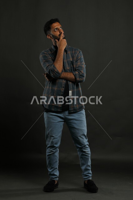 التأمل والتخطيط للمستقبل، إيجاد حل لمشكلة ما، بورتريه لشاب عربي خليجي سعودي يرتدي الزي الكاجوال يضع يده على ذقنه و ينظر إلى شيء ما بإيماءات التفكير، التمعن والتركيز وشرود الذهن ، صورة الجسم بالطول الكامل، خلفية سوداء
