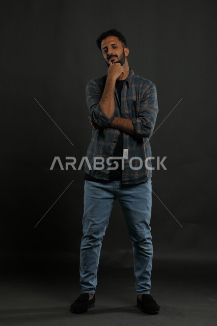 Finding a solution to a problem, contemplating and planning for the future, portrait of a Saudi Gulf Arab young man wearing casual clothes, placing his hand on his chin and looking at the camera with gestures of thought, contemplation, concentration and absent-mindedness, full-length body portrait, black background