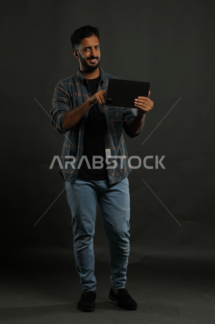 النظر إلى الكاميرا بإيماءات الذكاء، مشاهدة أخبار سارة على وسائل التواصل الاجتماعي، احتراف الوظائف التقنية وتكنولوجيا المعلومات، بورتريه لشاب عربي سعودي خليجي يرتدي اللباس الكاجوال يستخدم جهاز التابليت بإيماءات الفرح والسعادة, صورة الجسم بالكامل, خلفية سوداء