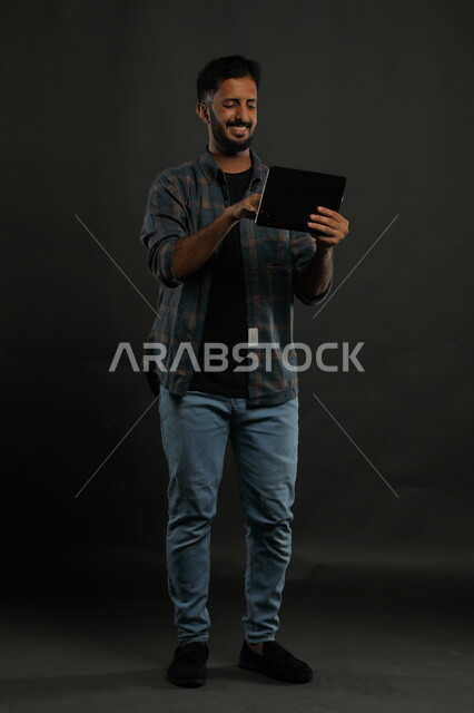 مشاهدة أخبار سارة على وسائل التواصل الاجتماعي، احتراف الوظائف التقنية وتكنولوجيا المعلومات، بورتريه لشاب عربي سعودي خليجي يرتدي اللباس الكاجوال يستخدم جهاز التابليت بإيماءات الفرح والسعادة, صورة الجسم بالكامل, خلفية سوداء