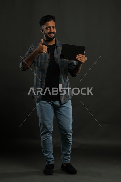 الشعور بالرضا والقبول بتقنيات الجوال, بورتريه لرجل عربي سعودي خليجي يرتدي الثوب والشماغ التقليدي مبتسم يستخدم الهاتف المحمول يرفع إصبعه نحو الأعلى بعلامة الجودة, الوقوف باستقامة والنظر إلى الكاميرا, صورة الجسم بالكامل, خلفية سوداء.