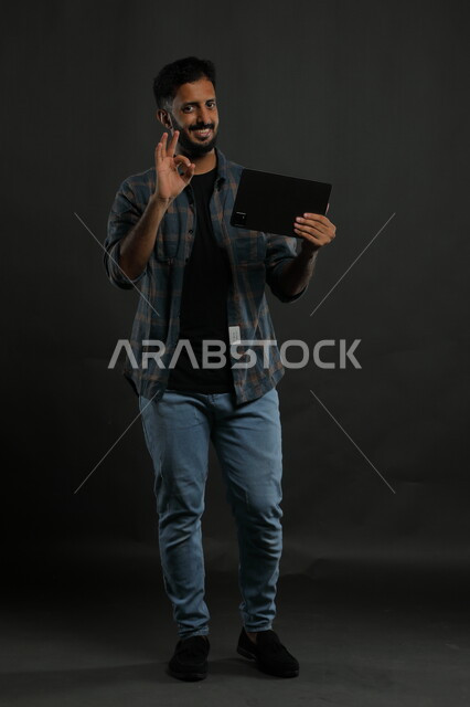 الإعجاب بآداء التقنيات الحديثة, بورتريه لشاب عربي سعودي خليجي يرتدي الثوب والشماغ التقليدي مبتسم يستخدم الهاتف المحمول ينظر إلى الكاميرا, إيماءات جودة الأجهزة المتطورة وتطبيقاتها, صورة الجسم بالكامل, خلفية سوداء.