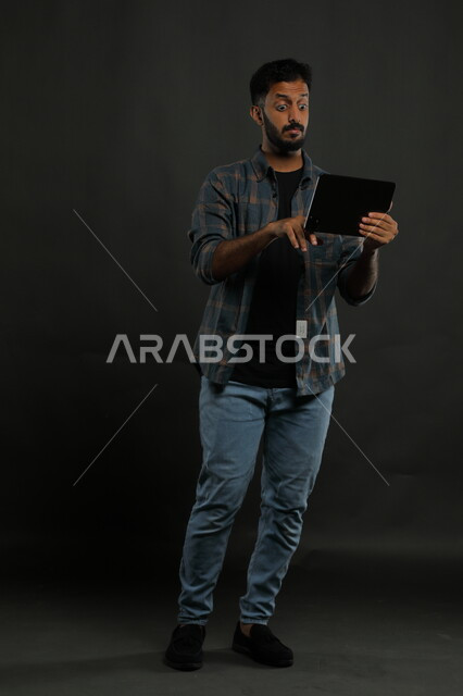 المفاجأة والدهشه بأمر ما، ايماءات الانفعال والاندماج أثناء مشاهدة  شيء ما على الجهاز اللوحي، بورتريه لرجل عربي خليجي سعودي يرتدي الزي الكاجوال يقوم بإجراء مكالمة فيديو وسماع خبر مفاجئ ،صورة الجسم بالطول الكامل، خلفية سوداء