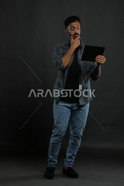 ايماءات الانفعال والاندماج أثناء مشاهدة  شيء ما على الجهاز اللوحي، المفاجأة والدهشه بأمر ما، بورتريه لرجل عربي خليجي سعودي يرتدي الزي الكاجوال يقوم بإجراء مكالمة فيديو وسماع خبر مفاجئ ،صورة الجسم بالطول الكامل، خلفية سوداء