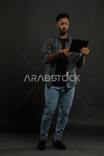 ايماءات الذهول والانفعال أثناء مشاهدة  شيء ما على الجهاز اللوحي، الانزعاج والغضب لأمر ما، بورتريه لرجل عربي خليجي سعودي يرتدي الزي الكاجوال يقف باستقامة وينقر على شاشة التابليت ،صورة الجسم بالطول الكامل، خلفية سوداء