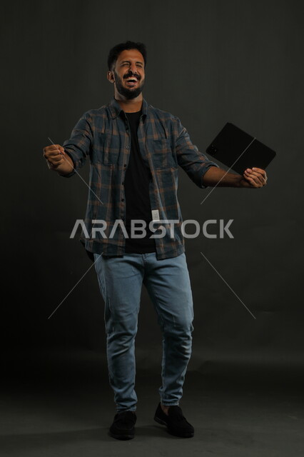 ميزات تقنية حديثة ومتطورة, الشعور بالحماس للنجاح وتحقيق الأهداف, بورتريه لشاب عربي سعودي خليجي يرتدي الزي الكاجوال يمسك التابليت يرفع يديه بإيماءات الفرح الشديد, الشعور بالاندهاش والذهول, صورة الجسم بالكامل, خلفية سوداء.