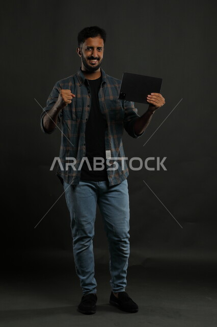 الشعور بالحماس للنجاح وتحقيق الأهداف, ميزات تقنية حديثة ومتطورة, بورتريه لشاب عربي سعودي خليجي يرتدي الزي الكاجوال يمسك التابليت يرفع يديه بإيماءات الفرح الشديد, السرور والفرح بالإنجازات, صورة الجسم بالكامل, خلفية سوداء.