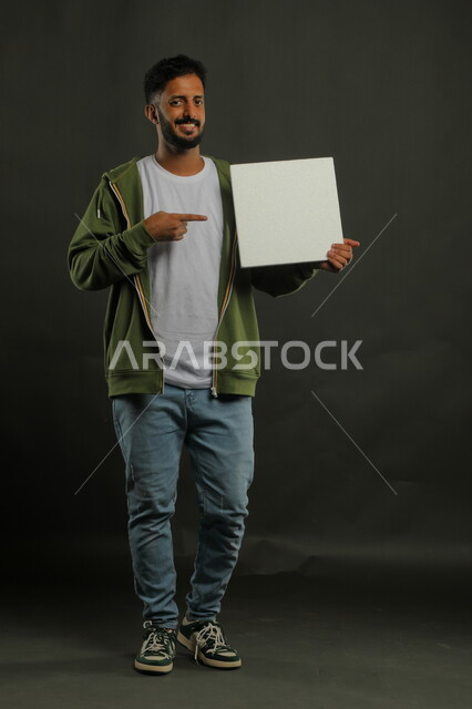 إعلانات وعروض تسويقية، موك اب مربع أبيض، استخدام لوحات توضيحية ووسائل تعليمية، بورتريه لشاب عربي خليجي سعودي مبتسم يرتدي الزي الكاجوال يمسك بيده لوحه بيضاء مربعه فارغة وينظر إلى الكاميرا، صورة الجسم بالكامل، خلفية سوداء
