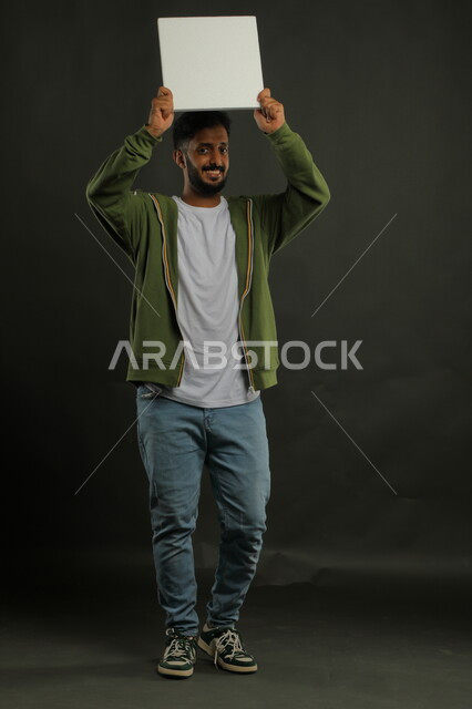 النظر إلى الكاميرا بإيماءات الثقة، إعلانات وعروض تسويقية، موك اب مربع أبيض، استخدام لوحات توضيحية ووسائل تعليمية، بورتريه لشاب عربي خليجي سعودي مبتسم يرتدي الزي الكاجوال يمسك بيده لوحه بيضاء مربعه فارغة يرفعها عاليا، صورة الجسم بالكامل، خلفية سوداء