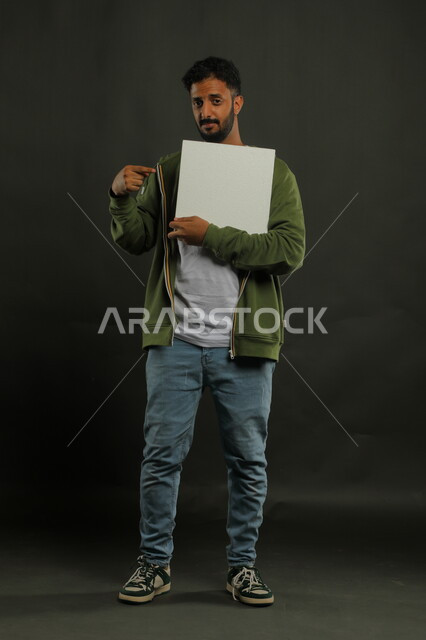 استخدام لوحات توضيحية ووسائل تعليمية، النظر إلى الكاميرا بإيماءات الثقة،إعلانات وعروض تسويقية، موك اب مربع أبيض، بورتريه لشاب عربي خليجي سعودي مبتسم يرتدي الزي الكاجوال يمسك بيده لوحه بيضاء مربعه فارغة، صورة الجسم بالكامل، خلفية سوداء