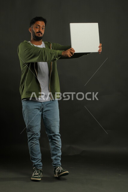 إعلانات وعروض تسويقية، موك اب مربع أبيض، استخدام لوحات توضيحية ووسائل تعليمية، بورتريه لشاب عربي خليجي سعودي مبتسم يرتدي الزي الكاجوال يمسك بيده لوحه بيضاء مربعه فارغة وينظر إلى الكاميرا، صورة الجسم بالكامل، خلفية سوداء