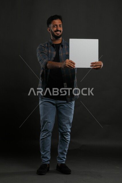 عروض وإعلانات تسويقية، موك اب مربع أبيض، استخدام لوحات توضيحية ووسائل تعليمية، بورتريه لشاب عربي خليجي سعودي مبتسم يرتدي الزي الكاجوال يمسك بيده لوحه بيضاء مربعه فارغة وينظر إلى الكاميرا، صورة الجسم بالكامل، خلفية سوداء
