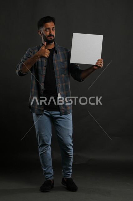 إعلانات وعروض تسويقية، موك اب مربع أبيض، استخدام لوحات توضيحية ووسائل تعليمية، بورتريه لشاب عربي خليجي سعودي مبتسم يرتدي الزي الكاجوال يمسك بيده لوحه بيضاء مربعه فارغة وينظر إلى الكاميرا، صورة الجسم بالكامل، خلفية سوداء